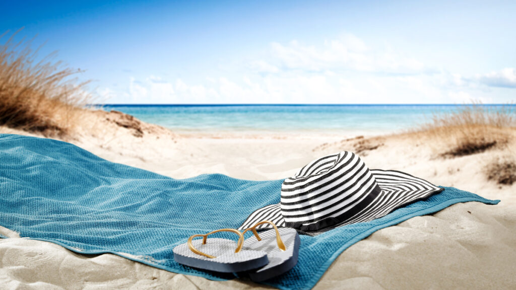 Serviette bleue, chapeau rayé et tongs sur une dune de sable face à l'océan azur.